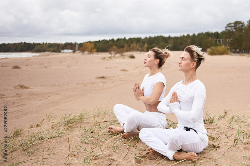 Yoga, zen, enlightenment, reacreation, meditation and concentration concept. Young male and female wearing white clothes meditating with eyes closed, making namaste gesture, sitting in lotus pose