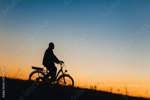 Male cyclist on the e-bike or electric bicycle on the sunset background slides down the hill. Silhouette of the old man in profile. Active pension. Travel. Sport.