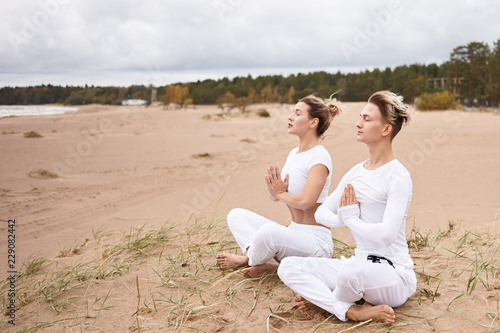 Yoga, zen, enlightenment, reacreation, meditation and concentration concept. Young male and female wearing white clothes meditating with eyes closed, making namaste gesture, sitting in lotus pose