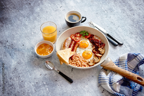 Full English Breakfast served in a pan. Fried eggs, beked beans, tomatoes, champignons, crispy bacon, sausages and toast. Placed on stone background. Top view with copy space.