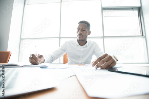 Serious busy African-American-American male manager in white shirt sitting at table full of papers and making notes in diary while doing business analysis in office.