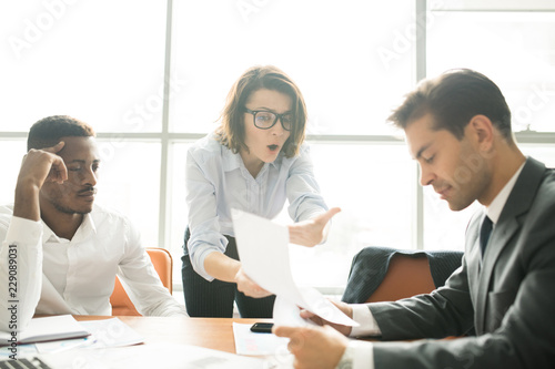 Shocked attractive young business lady in glasses showing report to colleague while proving her argument at staff meeting, indifferent businessman reading contract