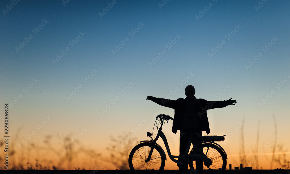 Obraz premium Silhouette of the male cyclist on the e-bike or electric bicycle on the sunset background. The man spread his arms to the side. Active pension. Relaxation. Travel. Sport.