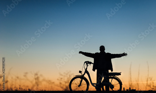 Silhouette of the male cyclist on the e-bike or electric bicycle on the sunset background. The man spread his arms to the side. Active pension. Relaxation. Travel. Sport.