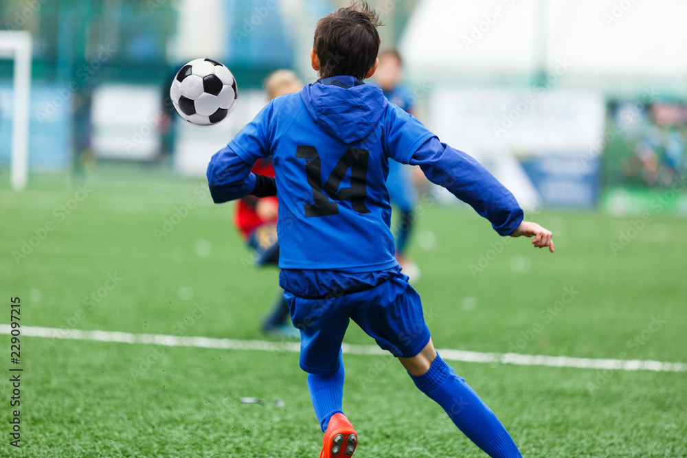 Children are kicking soccer classic white and black ball. Young kids football action. Boys are running after the Ball on green artificial grass. Footballers in blue and red shirts