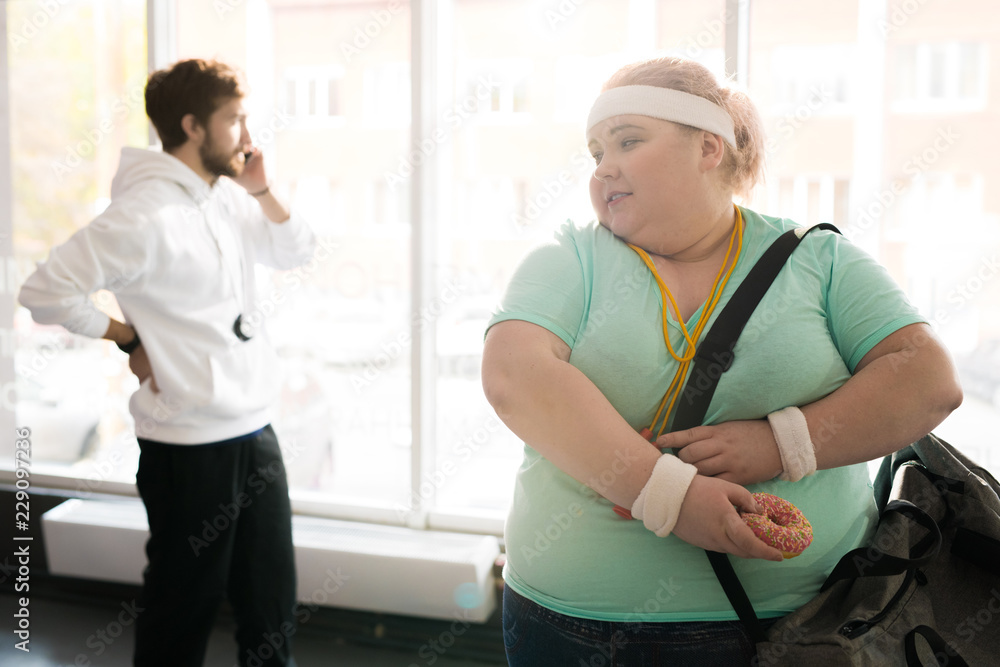 Waist up portrait of obese young woman holding donut leaving fitness training, copy space