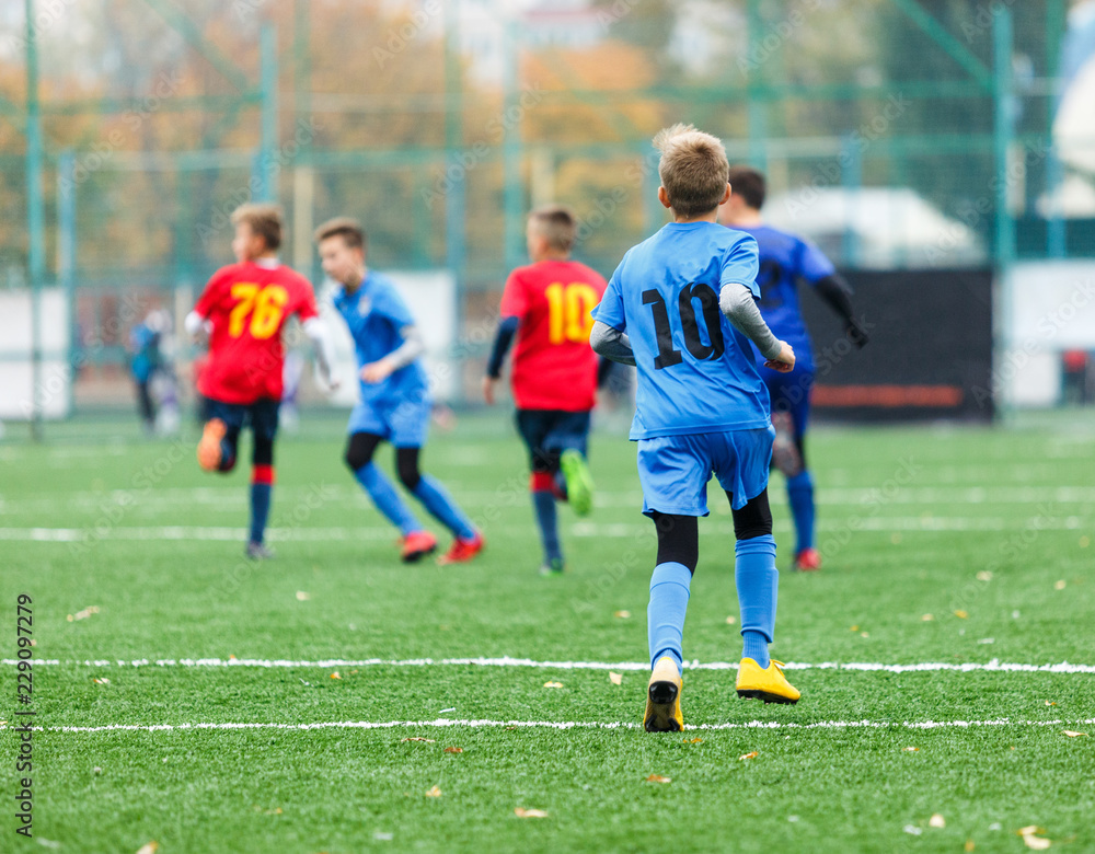 Children are kicking soccer classic white and black ball. Young kids football action. Boys are running after the Ball on green artificial grass. Footballers in blue and red shirts Children are kicking soccer classic white and black ball. Young kids football action. Boys are running after the Ball on green artificial grass. Footballers in blue and red shirts