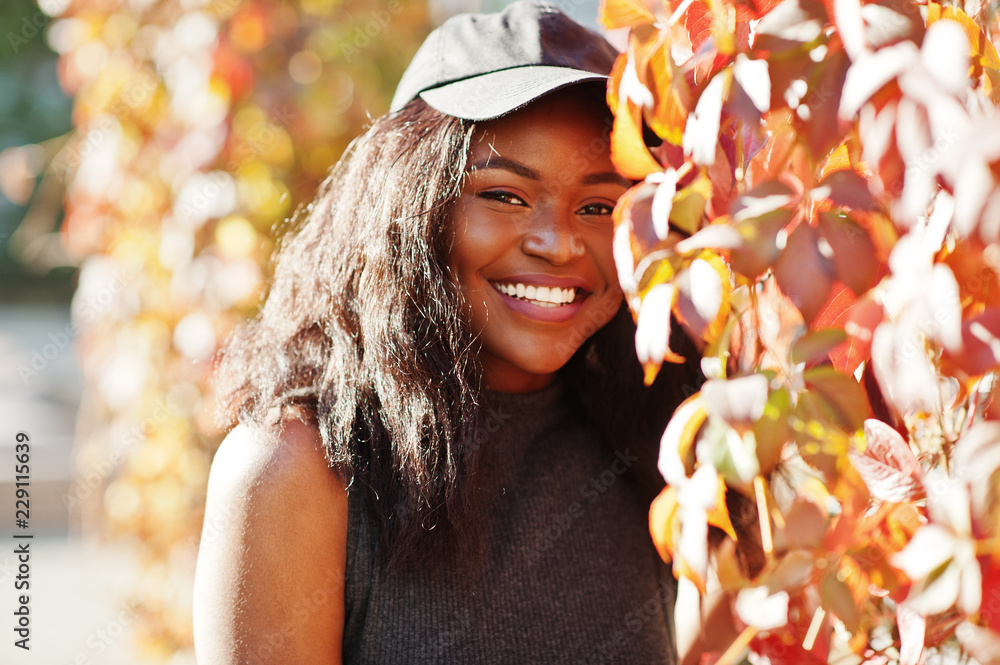 Fototapeta premium Stylish african american girl in cap posed at sunny autumn day against red leaves. Africa model woman.
