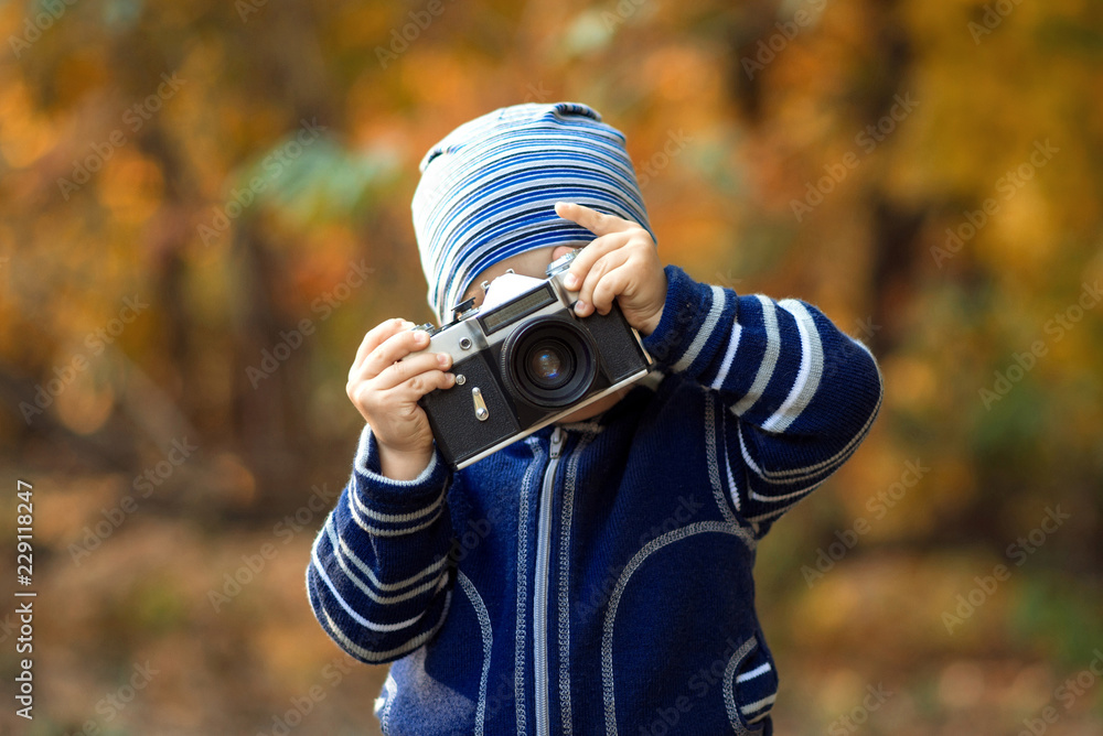 Portrait of a little child taking a picture with big vintage photo camera outdoor in autumn forest or park. Brown and golden trees in the background Portrait of a little child taking a picture with big vintage photo camera outdoor in autumn forest or park. Brown and golden trees in the background