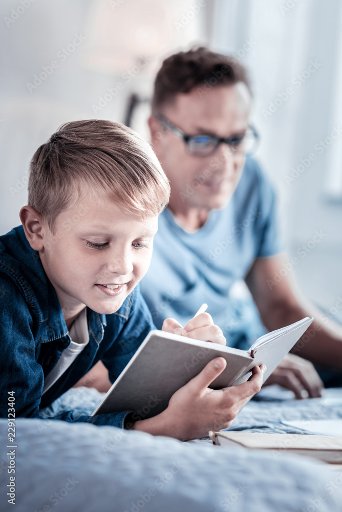 Correct the mistake. Concentrated pleasant little boy lying on the bed holding the notebook and writing. Correct the mistake. Concentrated pleasant little boy lying on the bed holding the notebook and writing.