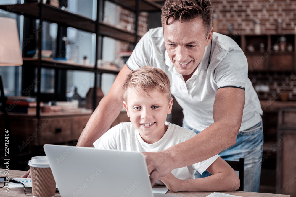 Pay your attention. Caring attentive handsome father standing near a boy who using the laptop pointing at the screen and giving advices to his son. Pay your attention. Caring attentive handsome father standing near a boy who using the laptop pointing at the screen and giving advices to his son.