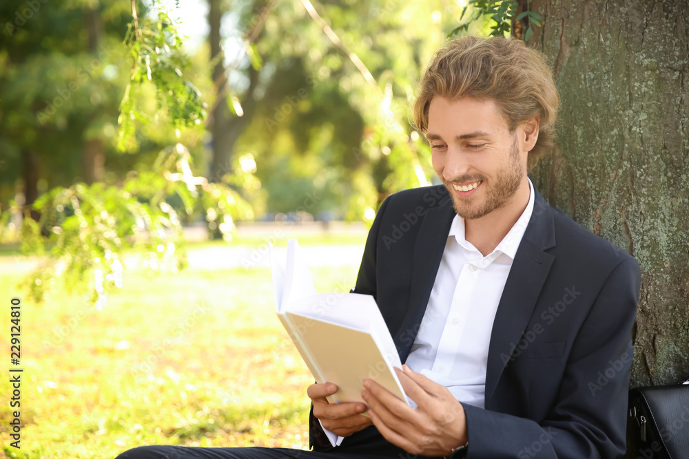 Handsome young businessman with book resting near tree in park Handsome young businessman with book resting near tree in park