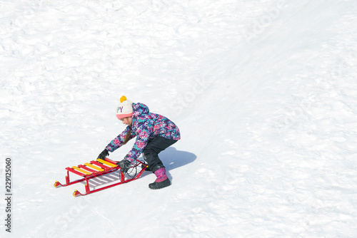 Cute little girl sliding on a sled on snowy hill. Active leisure outdoors in winter