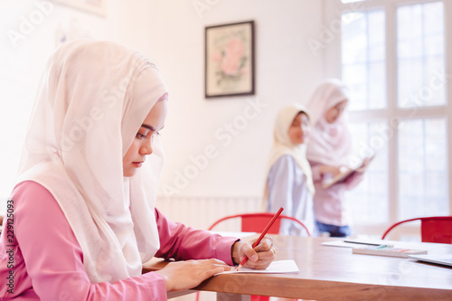 Beautiful young muslim teacher and cute asian muslim girls standing and reading a book in classroom.