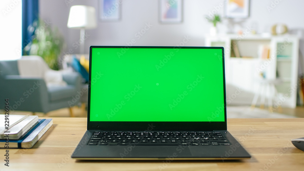 Modern Laptop with Green Mock-up Screen Display Standing on the Desk in the Cozy Living Room. Man with Mobile Phone Walks Through His Flat. Modern Laptop with Green Mock-up Screen Display Standing on the Desk in the Cozy Living Room. Man with Mobile Phone Walks Through His Flat.