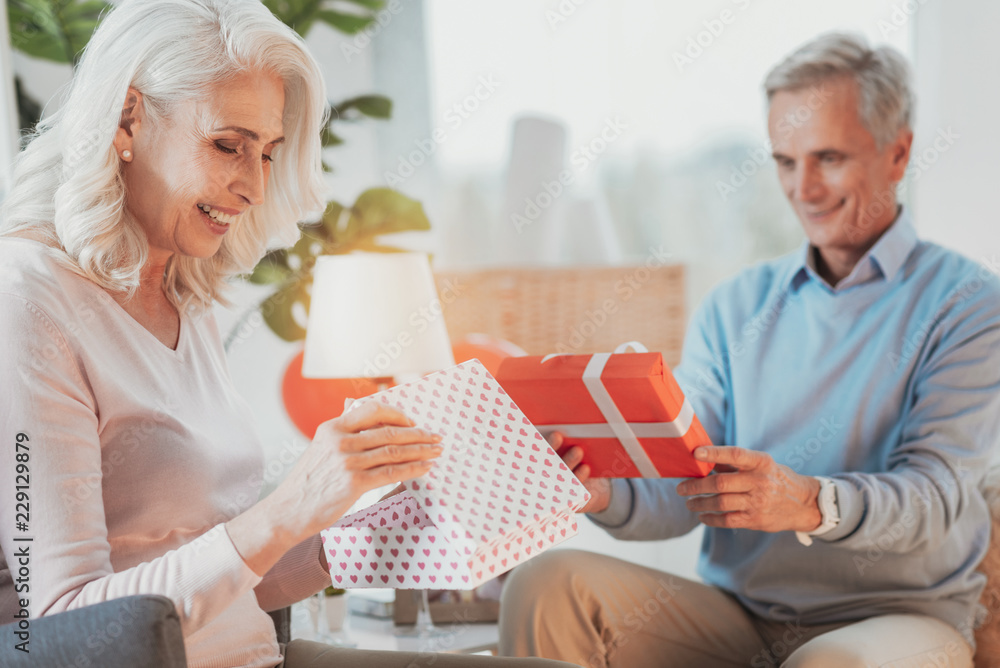 What is there. Happy elderly couple opening presented gifts to each other while relaxing in bright room and having brilliant mood