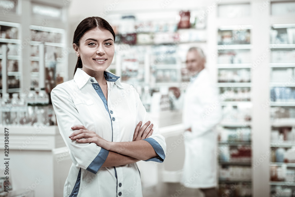 Woman in drugstore. Dark-haired beaming appealing woman wearing white coat working in big drugstore Woman in drugstore. Dark-haired beaming appealing woman wearing white coat working in big drugstore