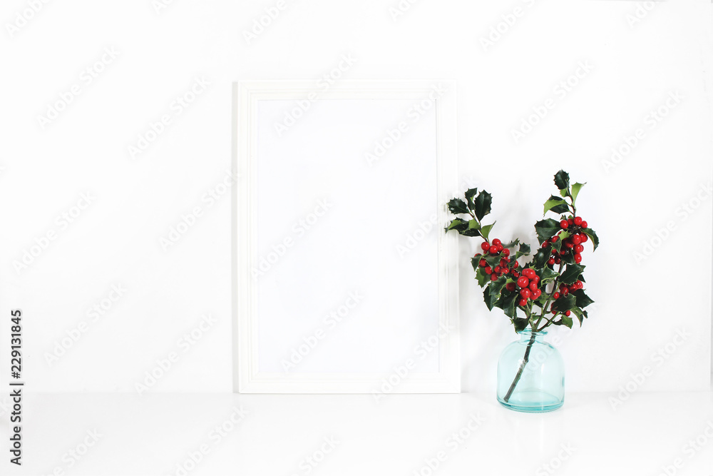 Fototapeta premium Vertical white blank wooden frame mockup. Holly berry branches in blue glass vase on white table. Styled stock feminine photography. Home decor. Christmas winter concept.