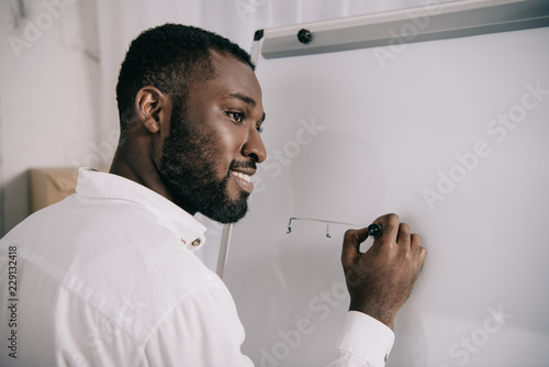 smiling handsome african american businessman drawing on flipchart in office and looking away