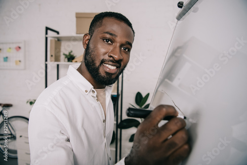 smiling handsome african american businessman drawing on flipchart in office and looking at camera