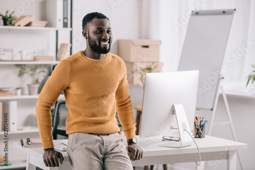 cheerful handsome african american designer in orange sweater leaning on table and looking away in office