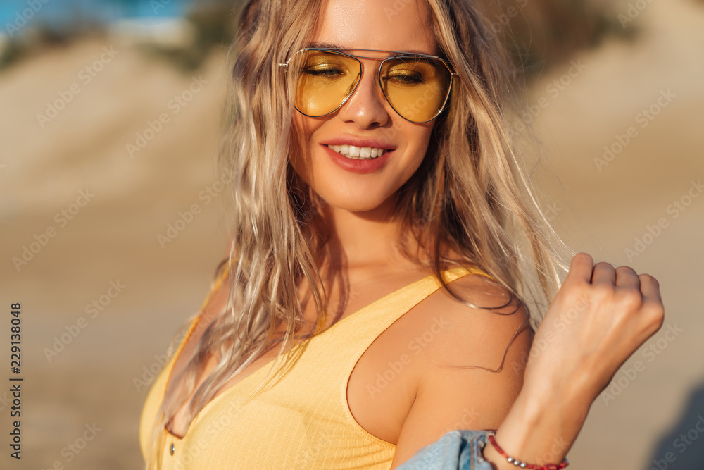 Close-up portrait of a sexy girl in a yellow swimsuit and sunglasses on the beach. Amazing woman face with make-up and beautiful hairstyle. Model on vacation, travels and sunbathes. Close-up portrait of a sexy girl in a yellow swimsuit and sunglasses on the beach. Amazing woman face with make-up and beautiful hairstyle. Model on vacation, travels and sunbathes.
