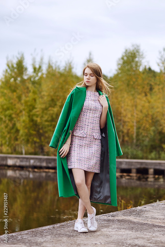 Girl in a green coat walks along the lake embankment on a cloudy autumn day. Autumn fashion and clothing, yellow fallen leaves floating in the water. Romantic mood
