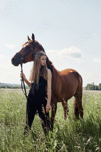 Girl rider stands next to the horse in the field. Fashion portrait of a woman and the mares are horses in the village in the grass. Blonde woman holding a horse by the bridle, beautiful body © angel_nt