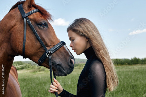 Girl rider stands next to the horse in the field. Fashion portrait of a woman and the mares are horses in the village in the grass. Blonde woman holding a horse by the bridle, beautiful body