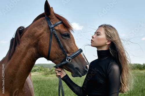 Girl rider stands next to the horse in the field. Fashion portrait of a woman and the mares are horses in the village in the grass. Blonde woman holding a horse by the bridle, beautiful body
