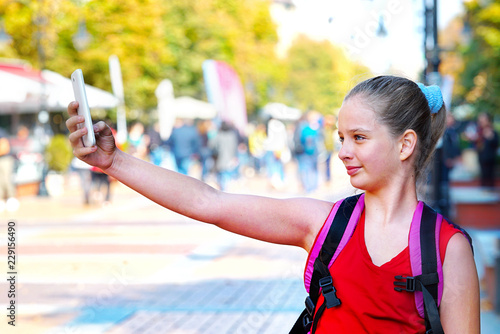 Tourist child girl with backpack outstretched arm by taking selfies on smartphone at cultural city pedestrianized street outdoor.