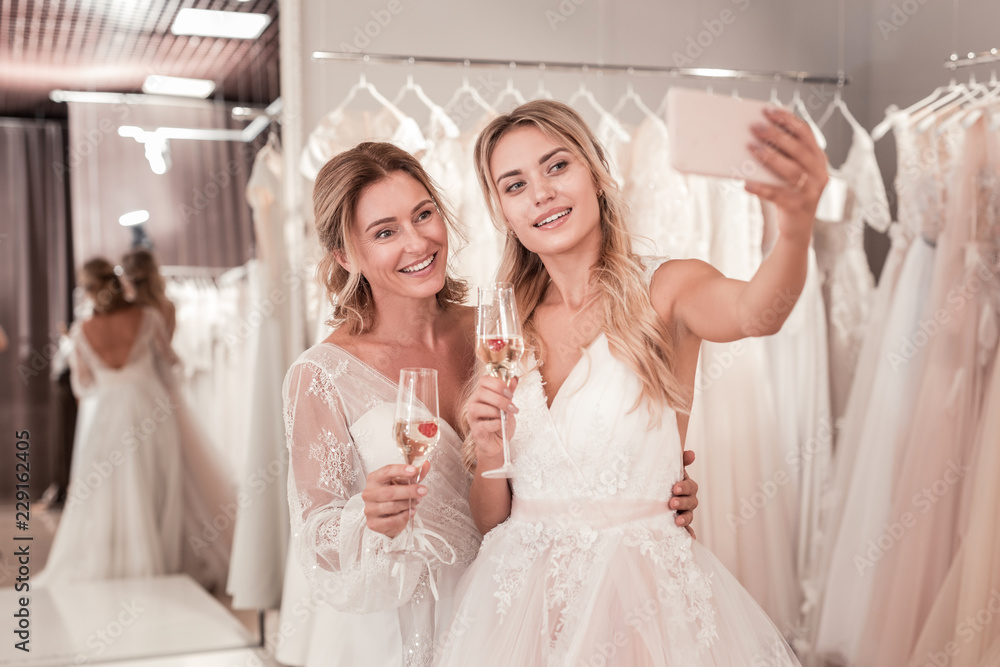 Beautiful photo. Joyful positive brides holding glasses with sparkling wine while taking a selfie together