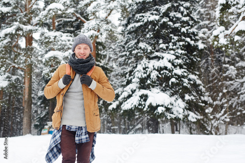 Smiling handsome young guy in knitted hat and scarf carrying backpack going down hill while hiking in winter forest