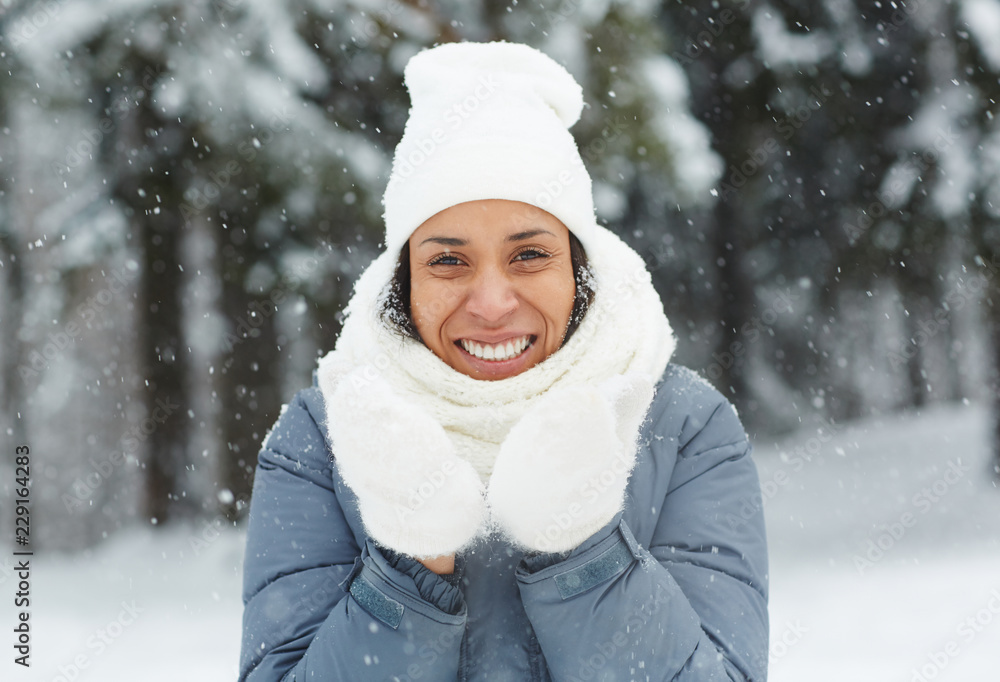 Obraz premium Cheerful excited attractive young woman in mittens wearing white hat and scarf looking at camera and standing under snow in winter forest