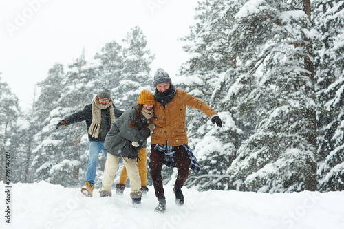 Positive excited young friends in warm clothing running over winter hill while having fun in forest