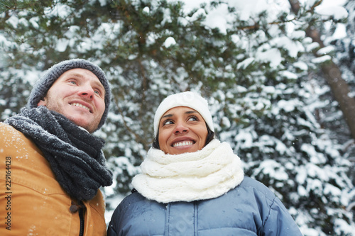 Smiling positive young multi-ethnic couple in warm hats and scarf looking around and enjoying beauty of nature while walking in winter park and being on vacation © pressmaster