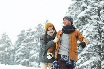 Jolly beautiful young couple in puffy jackets holding hands and laughing while enjoying winter stroll, hiking in forest