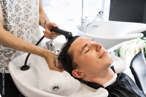 Relaxed young man with eyes closed leaning on sink while hairdresser washing his hair in salon