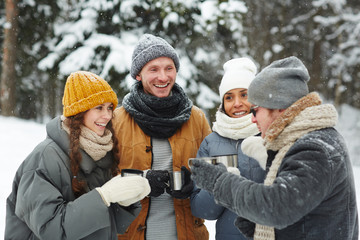 Happy excited young travelers in winterwear standing in forest and drinking hot tea while chatting at winter hike