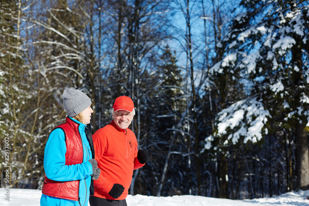 Mature spouses in activewear having talk while jogging in winter forest or park