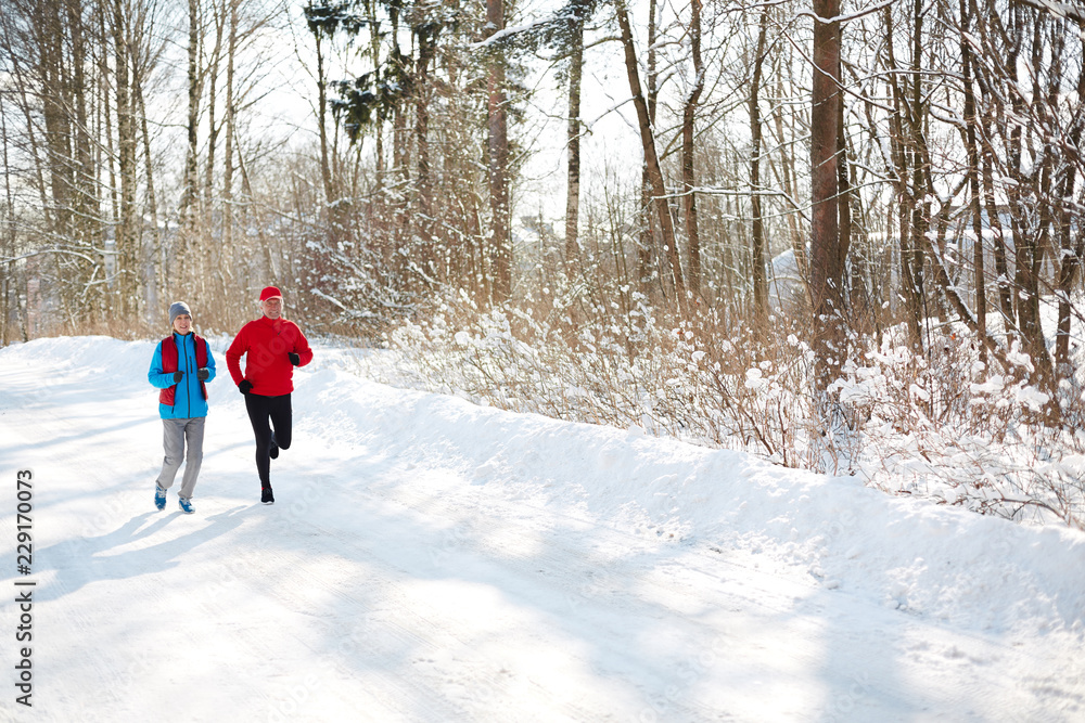 Mature couple in sportswear running down snow road in winter forest in the morning
