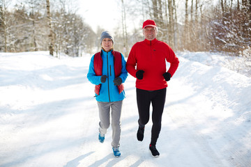 Active seniors jogging down winter road in the forest on sunny day or morning