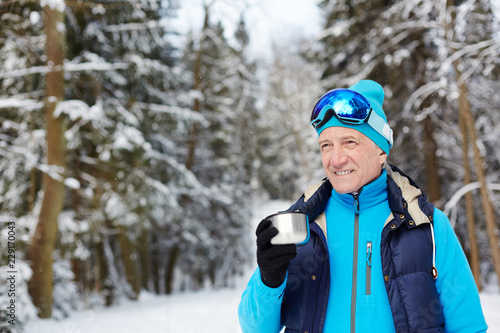 Active senior man in sportswear and goggles having hot tea in winter forest after training