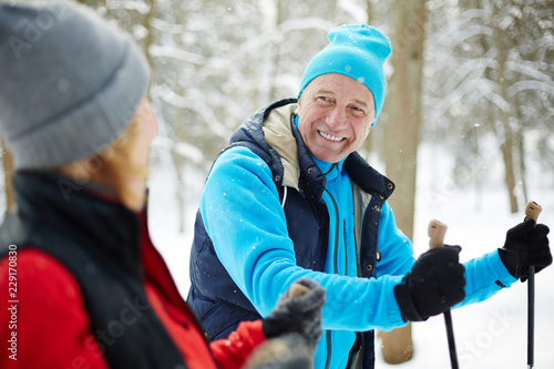 Happy mature man in activewear looking at his wife while skiing in winter forest at leisure