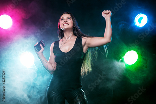 Beautiful young and woman in headphones listening music from phone and smiling. Color lights.