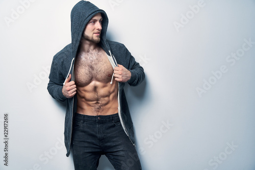 Brutal naked muscular man dressed in a grey hoodie posing by white wall looking to side at blank copy space