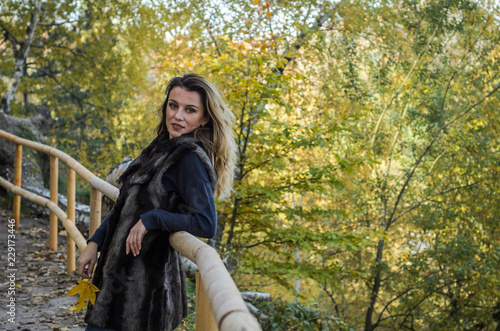 Young beautiful girl with long hair, in a fur jacket, stands on a wooden railing on a bright sunny day while walking in the autumn park