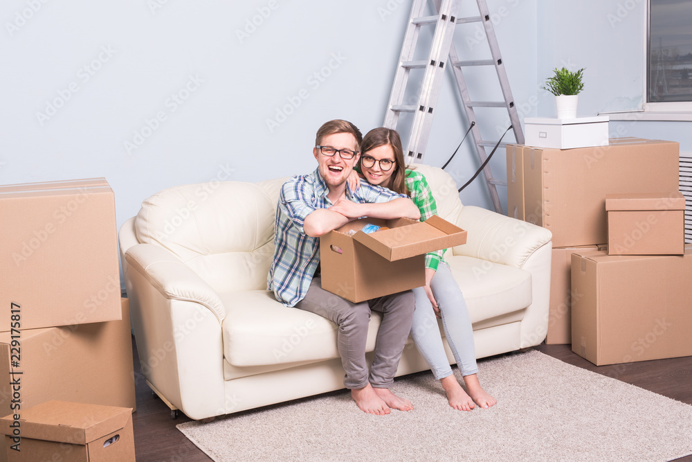 Home, relocation and moving concept - happy couple in glasses holding a card board and sitting on sofa Home, relocation and moving concept - happy couple in glasses holding a card board and sitting on sofa