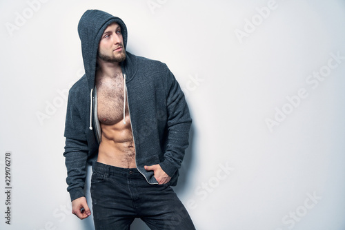 Fotografi Brutal naked muscular man dressed in a grey hoodie posing by white wall looking