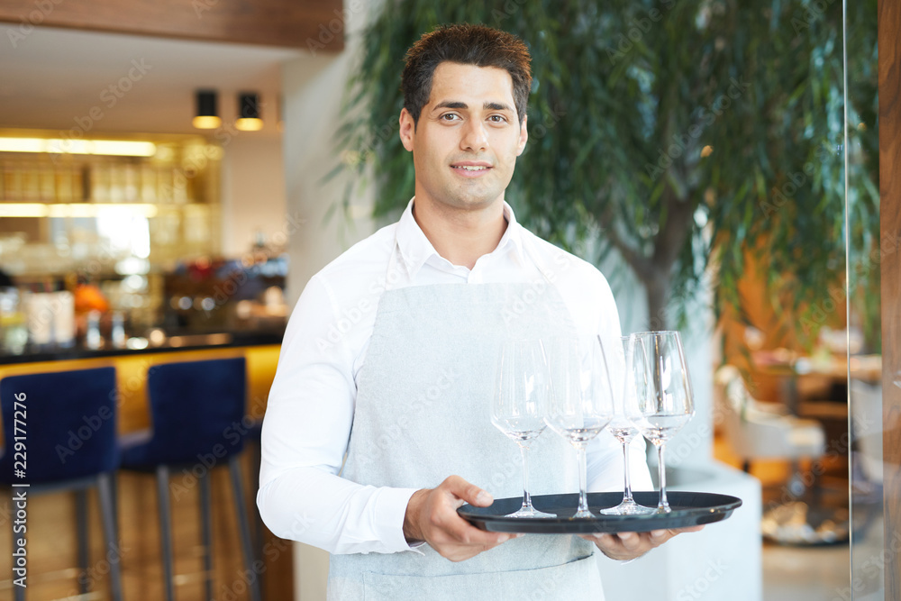 Portrait of young waiter in uniform holding tray with empty wineglasses standing at restaurant Portrait of young waiter in uniform holding tray with empty wineglasses standing at restaurant